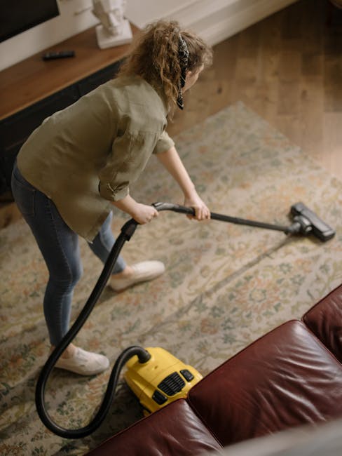 A yellow canister vacuum cleaner with a black hose is positioned on a patterned area rug in a living room, indicating surface cleaning and deep cleaning activity. The rug lies on a wooden floor with a