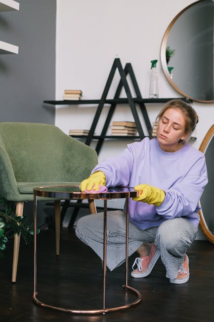A person wearing a dark jacket is performing surface cleaning on a decorative yellow and white patterned cushion placed on a dark fabric sofa. The individual holds a black vacuum cleaner attachment, w