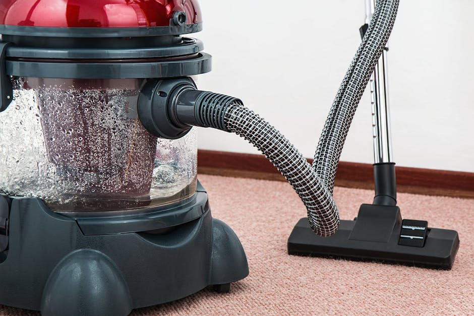 A professional cleaner dressed in a white protective suit, face mask, and blue gloves is performing deep carpet cleaning in a well-lit room. The cleaner is using a vacuum or steam cleaning machine to